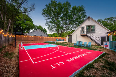 Residential backyard with bright red and blue multi-sport court and pickleball net, string lights along a wooden fence, Adirondack chairs, and a white two-story house with exterior stairs under a large tree at dusk.