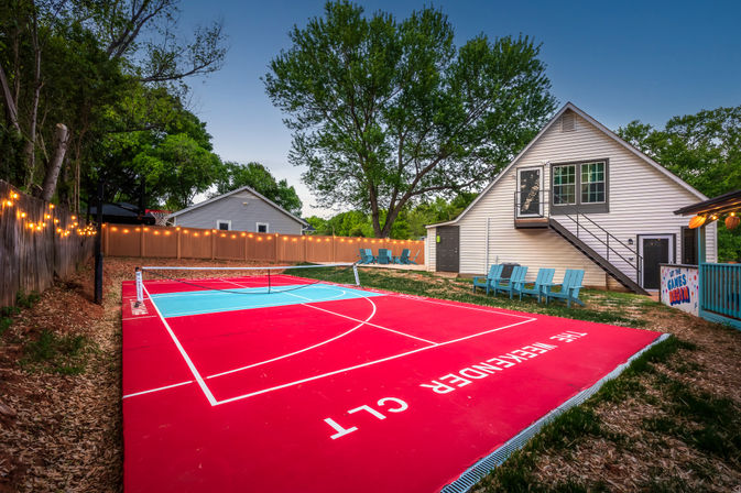 Residential backyard with bright red and blue multi-sport court and pickleball net, string lights along a wooden fence, Adirondack chairs, and a white two-story house with exterior stairs under a large tree at dusk.