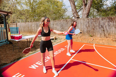 Two women playing pickleball on a bright red backyard court on a sunny day, holding paddles and a perforated ball, with a folding table of red cups and a giant Connect Four game by a wooden fence.