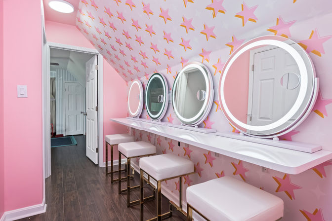 Playful pink beauty room with four round LED-lit vanity mirrors above a white floating counter, four white cushioned stools with gold legs, pink star-pattern wallpaper, dark wood floor and an open doorway.