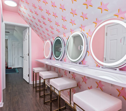 Playful pink beauty room with four round LED-lit vanity mirrors above a white floating counter, four white cushioned stools with gold legs, pink star-pattern wallpaper, dark wood floor and an open doorway.