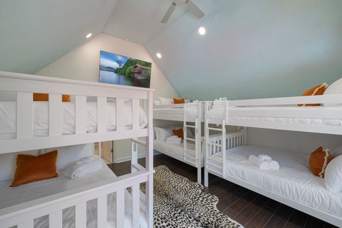 Cozy attic-style bunk room with vaulted ceiling, multiple white bunk beds dressed in crisp white linens, rust-orange throw pillows and rolled towels, dark hardwood floors with a leopard-print rug, ceiling fan and lakeside wall art.