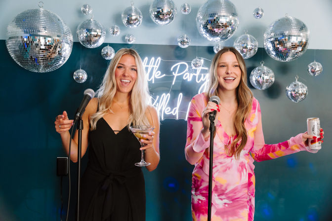 Two friends singing karaoke at a disco‑themed indoor party with hanging mirrored disco balls and a neon sign, one holding a martini glass and the other a canned drink.