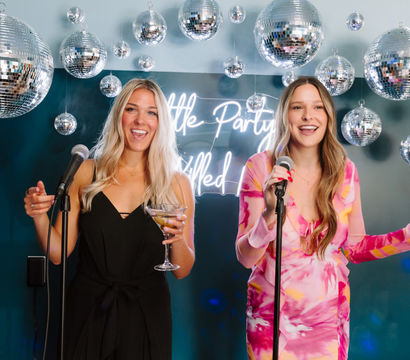 Two friends singing karaoke at a disco‑themed indoor party with hanging mirrored disco balls and a neon sign, one holding a martini glass and the other a canned drink.
