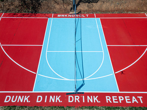 Aerial view of a colorful outdoor court with red outer sections and a blue center, a net down the middle casting a shadow, and white baseline text reading 'DUNK. DINK. DRINK. REPEAT.'