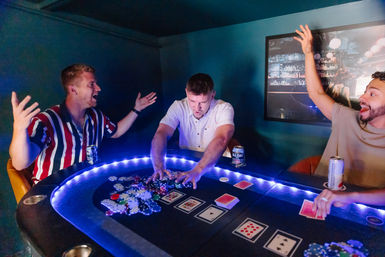 Three friends at a home poker night in a dim game room around an LED-lit poker table — center player sweeps a pile of chips while others cheer, cards and beer cans on the table.