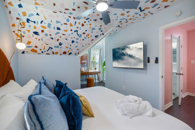 Cheerful attic bedroom with sloped terrazzo-patterned ceiling, pale blue walls, white bed with blue and gold pillows, ceiling fan, wall-mounted TV, desk by window and pink bathroom doorway.