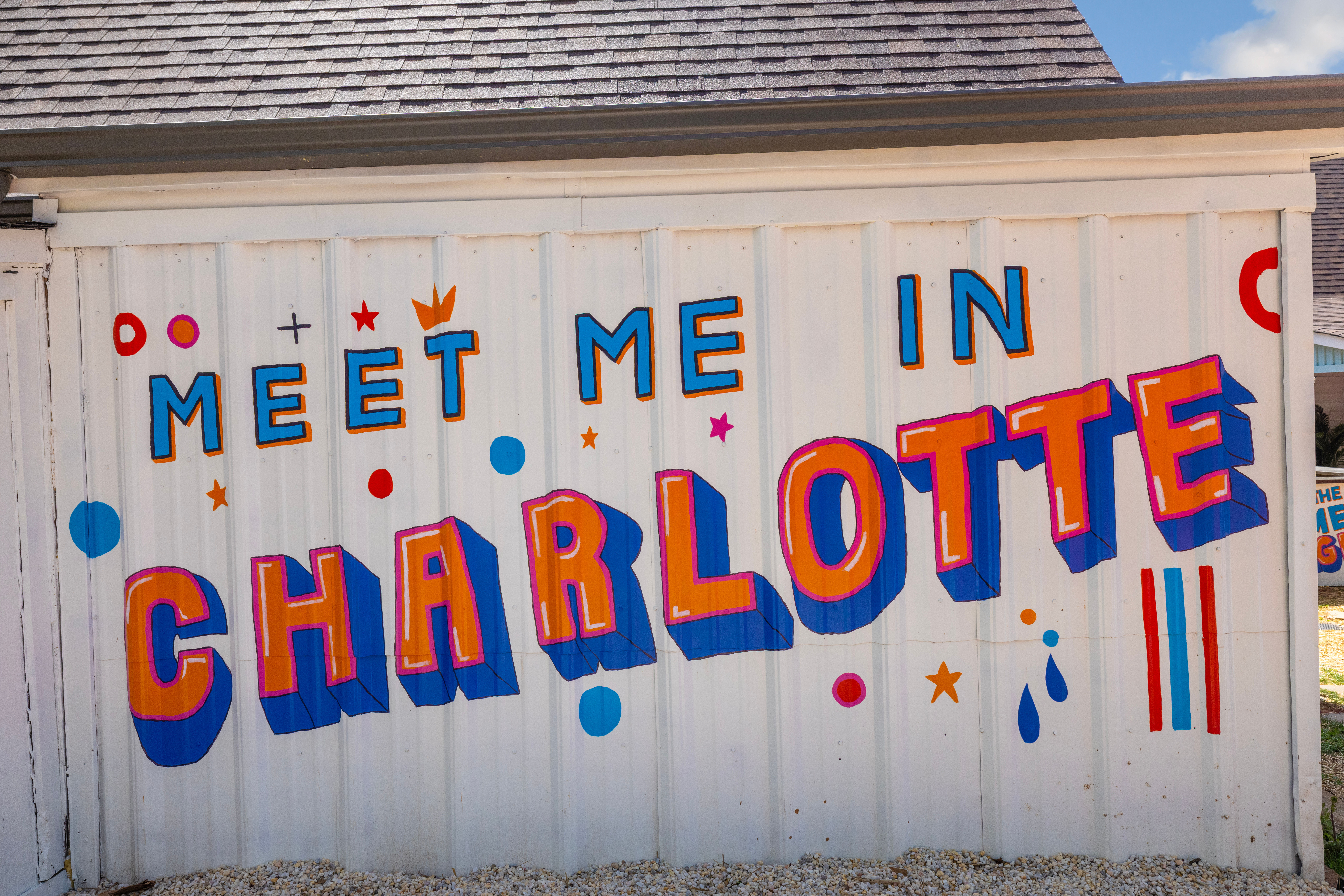 Playful outdoor mural reading 'MEET ME IN CHARLOTTE' on a white corrugated wall, featuring bold 3D orange and blue letters with colorful dots and stars.