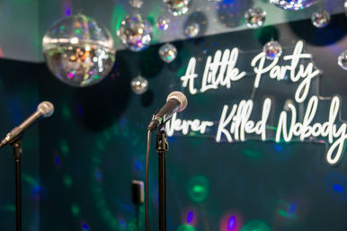 Two microphones on stands at an indoor karaoke/party stage with hanging disco balls, colorful light spots, and a neon sign reading “A Little Party Never Killed Nobody”.