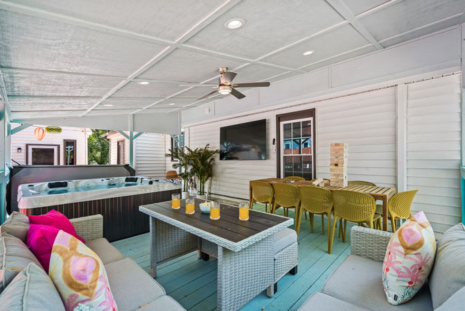 Covered backyard patio and outdoor living area with hot tub, wicker sectional with colorful pillows, coffee table with orange drinks, wall-mounted TV, ceiling fan, and long dining table with yellow chairs.