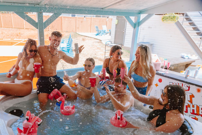 Young adults in swimsuits laughing and toasting with red cups and pink flamingo cup floats in a backyard hot tub under a covered patio, water splashing at a sunny summer party.