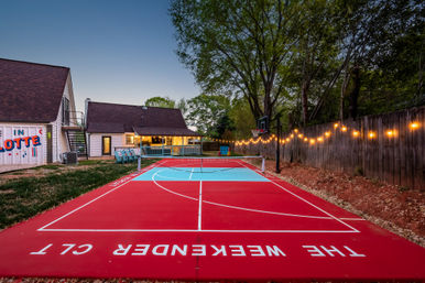 Vibrant red-and-turquoise backyard multi-sport court at dusk in Charlotte, with a basketball hoop, net, string lights along a wooden fence, and a cozy lit house with a porch in the background.