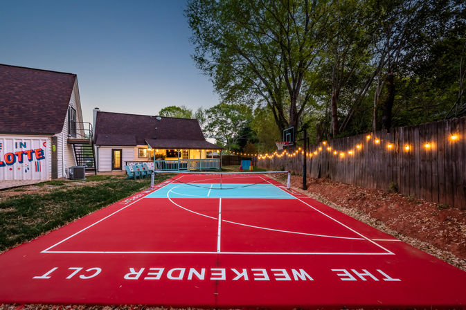 Vibrant red-and-turquoise backyard multi-sport court at dusk in Charlotte, with a basketball hoop, net, string lights along a wooden fence, and a cozy lit house with a porch in the background.