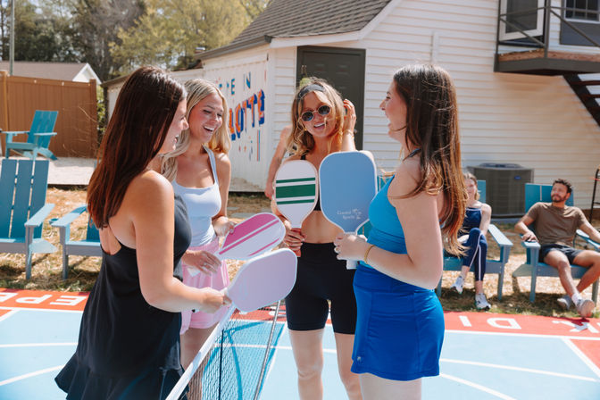 Four friends laughing over the net during a sunny backyard pickleball game, holding colorful paddles on a blue outdoor court with others relaxing in Adirondack chairs by a white house.