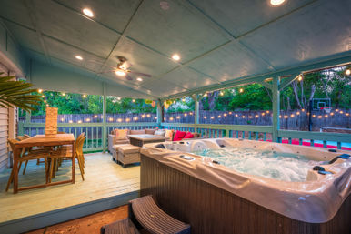Cozy covered backyard patio at dusk with a bubbling hot tub, wicker sectional with colorful cushions, wooden dining table with giant Jenga, string lights and ceiling fan overlooking a fenced suburban yard and basketball hoop.