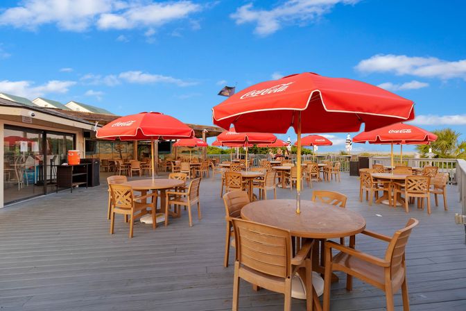 Sun-soaked beachside dining deck with round wooden tables and bright red umbrellas on a blue-sky day, overlooking coastal water and palm trees.
