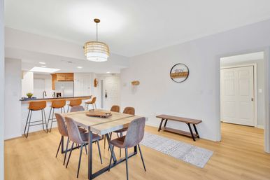 Bright modern open-plan dining area and kitchen with light wood floors, rectangular wood dining table and six brown chairs, breakfast bar with four tan leather stools, pendant light, stainless steel refrigerator and minimalist neutral decor.