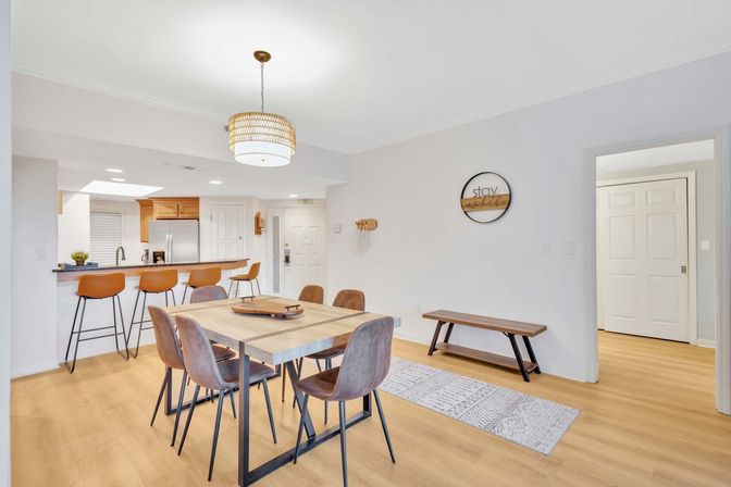 Bright modern open-plan dining area and kitchen with light wood floors, rectangular wood dining table and six brown chairs, breakfast bar with four tan leather stools, pendant light, stainless steel refrigerator and minimalist neutral decor.