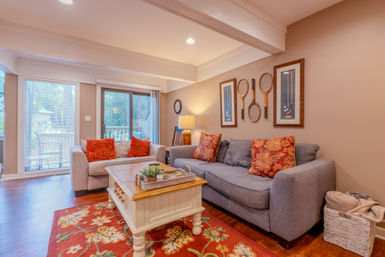 Cozy living room with gray sofa and beige loveseat, red floral rug, wooden coffee table, vintage tennis rackets on the wall and sliding glass door to a deck