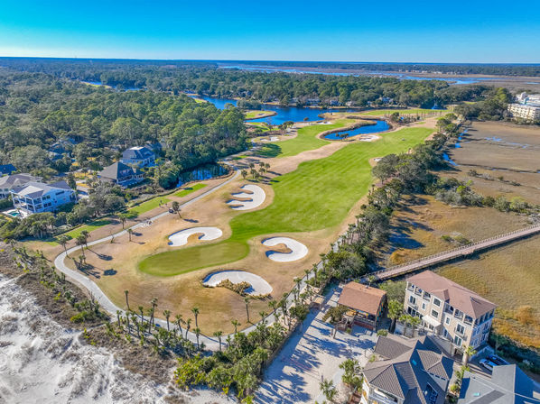 Sunny aerial view of an oceanfront coastal golf course with sculpted bunkers and green fairways winding past lagoons, marshland, beachfront homes and a boardwalk under a bright blue sky.