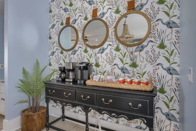 Home entryway coffee station with a black vintage console holding a Keurig and drip coffee maker, woven tray of snacks and apples, three round leather‑strapped mirrors, heron marsh botanical wallpaper and a potted fern