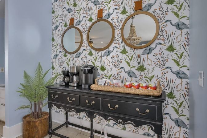 Home entryway coffee station with a black vintage console holding a Keurig and drip coffee maker, woven tray of snacks and apples, three round leather‑strapped mirrors, heron marsh botanical wallpaper and a potted fern