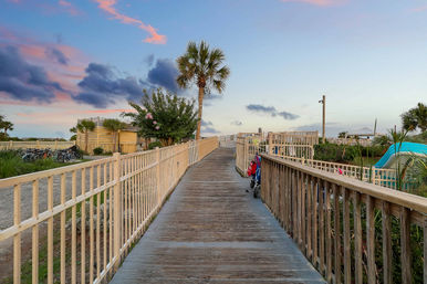 Wooden boardwalk leading to the beach, lined with palm trees and parked bikes under pink-and-blue sunset clouds