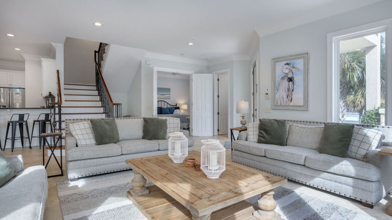 Sunlit coastal open-plan living room with two gray sofas and green pillows facing a rustic wooden coffee table, staircase to upper level, kitchen bar stools, coastal heron artwork, and large windows showing palm trees.