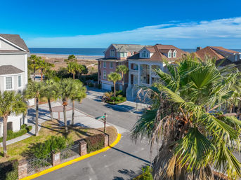 Aerial view of a sunny coastal neighborhood with pastel beach houses, palm trees lining a quiet street, and the sandy shoreline and blue ocean in the background.