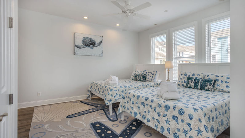 Bright coastal bedroom in a beach cottage with two beds dressed in blue seashell quilts, folded towels on each bed, sea turtle wall art, large sunlit windows with blinds, a bedside lamp, beach-themed rug and hardwood floor.