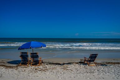 Sunlit sandy beach with two wooden lounge chairs under a bright blue umbrella and a lone chair facing gentle ocean waves and a clear blue sky