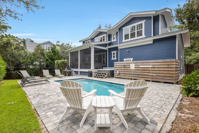 Backyard of a blue two-story suburban home featuring a rectangular swimming pool, two white Adirondack chairs with a small table on a paver patio, several lounge chairs, a screened porch and landscaped greenery.