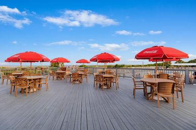 Sunny coastal boardwalk deck with wooden dining tables and bright red umbrellas overlooking sandy dunes and the ocean under a clear blue sky.