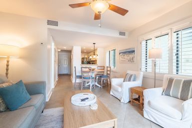 Sunlit open-concept coastal-style living room and dining area with ceiling fan, white slipcovered armchairs with striped pillows, teal-accent sofa, wooden coffee table, round dining table under pendant lights, tile floors and shuttered windows.