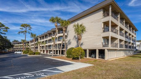 Sunlit three-story coastal condominium with private balconies, palm trees, reserved parking and bicycles stored beneath the raised ground floor under a bright blue sky.