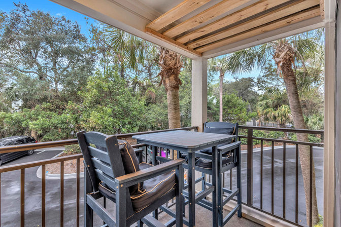 Covered balcony dining area with high wooden table and cushioned chairs overlooking palm trees, tropical foliage, and a parking area.