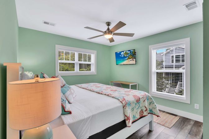 Sunny coastal-style bedroom interior with mint-green walls, large bed dressed in coral-and-aqua quilt, bedside lamp, modern ceiling fan, wall-mounted TV showing a tropical beach, two white-trim windows with blinds, small desk and wood-look flooring.