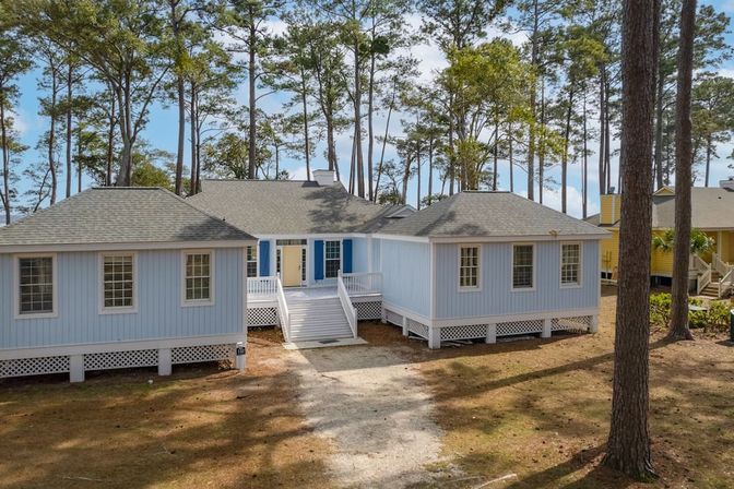 Light-blue elevated beach cottage with white stairs, yellow front door and blue shutters, nestled among tall pine trees with a sandy driveway on a sunny day