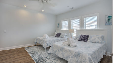 Bright coastal-style bedroom with two double beds, white headboards, blue floral quilts, navy accent pillows and folded towels on the beds, three windows with white shutters, bedside lamp and wood flooring.