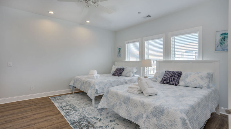 Bright coastal-style bedroom with two double beds, white headboards, blue floral quilts, navy accent pillows and folded towels on the beds, three windows with white shutters, bedside lamp and wood flooring.