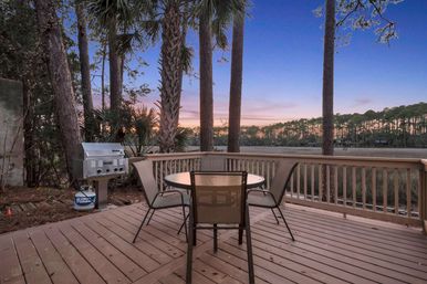Relaxing sunset waterfront deck with round table and four mesh chairs, stainless gas grill, palm and pine trees overlooking a coastal marsh and tree-lined horizon