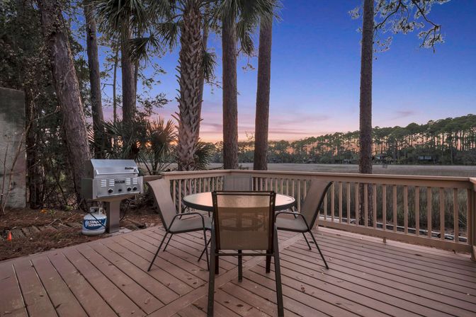 Relaxing sunset waterfront deck with round table and four mesh chairs, stainless gas grill, palm and pine trees overlooking a coastal marsh and tree-lined horizon