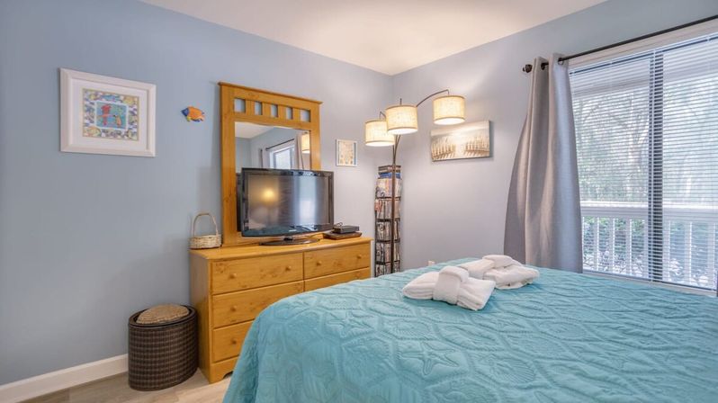 Cozy sunlit coastal-style bedroom with pale blue walls, wooden dresser and mirror topped by a flat-screen TV, aqua quilted bed with neatly folded white towels, arc floor lamp, and sliding glass door leading to a balcony.