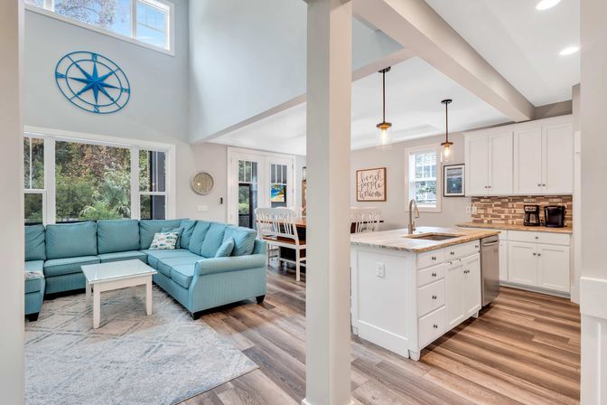 Sunlit open-concept coastal living room and kitchen with a teal L-shaped sofa, white marble-topped island, pendant lights, wood-look floors, compass wall decor and dining area.