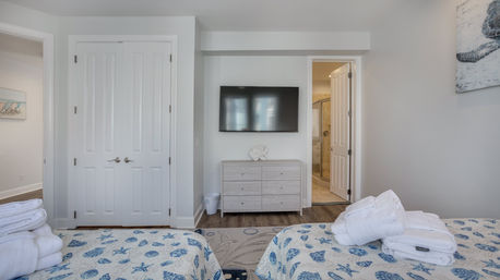 Bright coastal-themed twin bedroom in a vacation rental: two beds with seashell quilts, wall-mounted TV, light wood dresser, white double closet and en-suite bathroom.