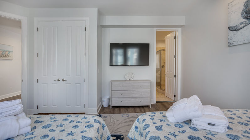 Bright coastal-themed twin bedroom in a vacation rental: two beds with seashell quilts, wall-mounted TV, light wood dresser, white double closet and en-suite bathroom.