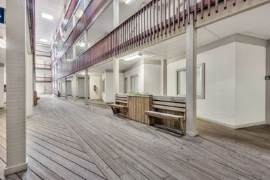 Bright multi-level apartment complex corridor with wood-plank deck flooring, upper balcony railings, built-in wooden benches and planter boxes under skylights