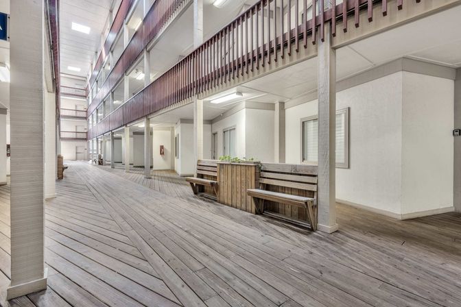 Bright multi-level apartment complex corridor with wood-plank deck flooring, upper balcony railings, built-in wooden benches and planter boxes under skylights