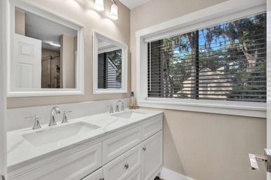 Sunlit modern master bathroom with white double-sink vanity, marble countertop and chrome faucets, two framed mirrors and a large window with horizontal blinds revealing leafy trees outside.