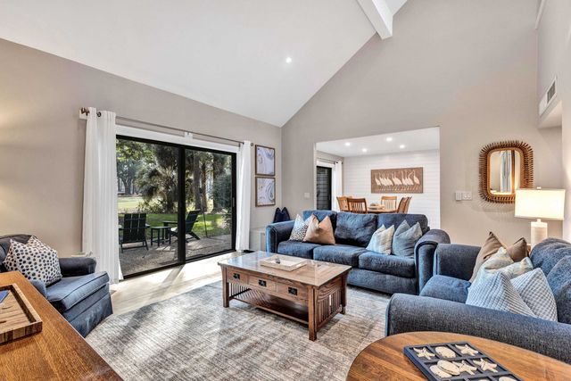 Sunlit coastal-inspired living room with vaulted ceiling, blue sofas, wooden coffee table and area rug, sliding glass doors opening to a leafy patio with outdoor chairs and a dining area visible in the background.